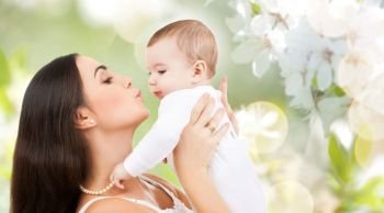 family and motherhood concept - happy smiling young mother kissing little baby over cherry blossom background. mother kissing baby over cherry blossom background