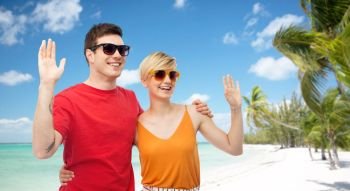 travel, tourism and summer holidays concept - smiling couple in sunglasses hugging and waving hands over exotic tropical beach with palm trees background. couple in sunglasses hugging and waving hands