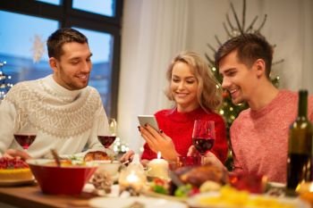 holidays and celebration concept - happy friends with smartphone having christmas dinner at home. friends with cellphone celebrate christmas at home
