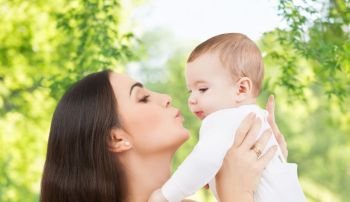 family and motherhood concept - happy smiling young mother kissing little baby over green natural background. mother kissing baby over green natural background
