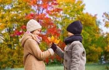 childhood, season and love concept - smiling little boy giving maple leaves to girl over autumn park background. kids with autumn maple leaves over park background