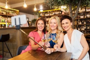 people, technology and lifestyle concept - women drinking wine and taking picture by smartphone on selfie stick at bar or restaurant. women taking picture by selfie stick at wine bar