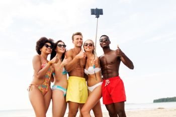 friendship, summer holidays and people concept - group of happy friends taking picture by smartphone on selfie stick on beach and showing thumbs up. friends taking selfie on beach and show thumbs up