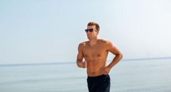 fitness, sport, people and healthy lifestyle concept - happy young man in sunglasses running along summer beach. happy man running along summer beach