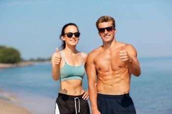fitness, sport and lifestyle concept - happy couple in sports clothes and sunglasses on beach showing thumbs up. couple of athletes showing thumbs up on beach