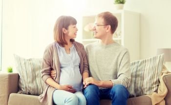 pregnancy, family and people concept - happy pregnant wife with husband sitting on sofa at home. happy pregnant wife with husband at home