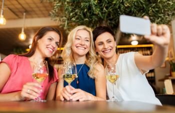 people, technology and lifestyle concept - women drinking wine and taking selfie by smartphone at bar or restaurant. women taking selfie by smartphone at wine bar