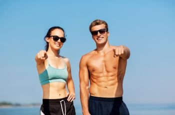 fitness, sport and lifestyle concept - happy couple in sports clothes and sunglasses on beach pointing finger to you. happy couple in sports clothes and shades on beach