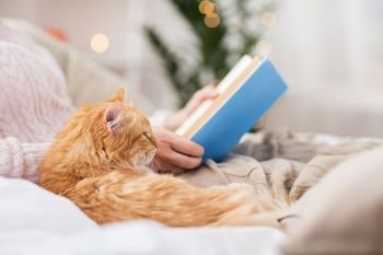 pets, hygge and people concept - red tabby cat and female owner reading book in bed at home. red cat and female owner reading book at home