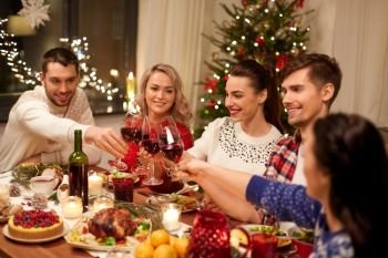holidays and celebration concept - happy friends having christmas dinner at home, drinking red wine and clinking glasses. friends celebrating christmas and drinking wine