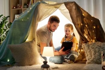 family, hygge and technology concept - happy father and little daughter with tablet pc computer in kids tent at night at home. family with tablet pc in kids tent at home