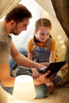 family, hygge and technology concept - happy father and little daughter with tablet pc computer in kids tent at night at home. family with tablet pc in kids tent at home