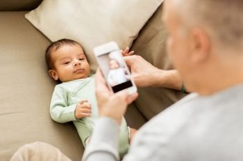 family, fatherhood and people concept - close up of father photographing little baby son by smartphone at home. father photographing baby by smartphone