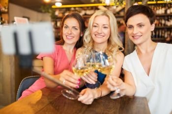 people, technology and lifestyle concept - women drinking wine and taking picture by smartphone on selfie stick  at bar or restaurant. women taking picture by selfie stick at wine bar