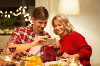 holidays, technology and celebration concept - happy couple having christmas dinner at home and photographing food by smartphone. couple with smartphone at home christmas dinner