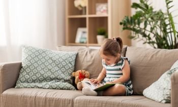 childhood and people concept - little girl sitting on sofa reading book and toy teddy bear at home. little girl reading book at home