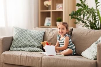childhood and people concept - happy little girl sitting on sofa with book and toy teddy bear at home. happy girl sitting on sofa with book at home