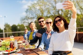 leisure and people concept - happy friends with drinks having dinner party on rooftop in summer and taking selfie by smartphone. happy friends taking selfie at rooftop party