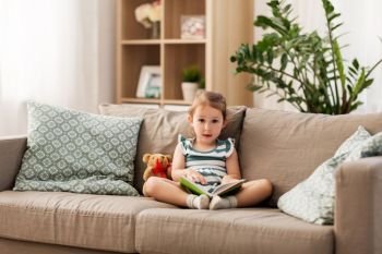 childhood and people concept - happy little girl sitting on sofa with book and toy teddy bear at home. happy girl sitting on sofa with book at home