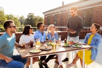 leisure and people concept - happy party host offering meat to his friends at barbecue party on rooftop in summer. friends at barbecue party on rooftop in summer