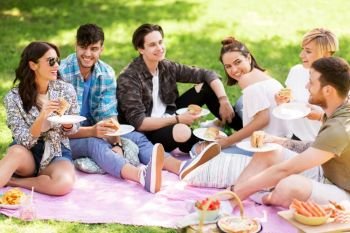 friendship, leisure and fast food concept - group of happy friends eating sandwiches or burgers at picnic in summer park. happy friends eating sandwiches at summer picnic