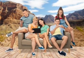 friendship, leisure and technology concept - group of happy smiling friends with tablet pc computer and smartphones sitting on sofa over grand canyon national park background. friends with tablet pc and smartphones sit on sofa