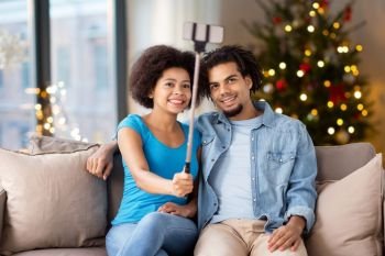 holidays, technology and people concept - happy african american couple with smartphone taking selfie at home over christmas tree lights background. couple with smartphone taking selfie on christmas