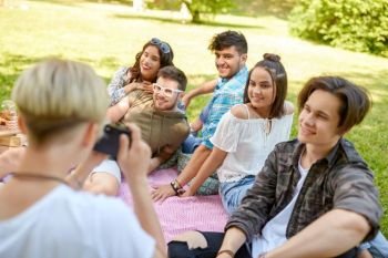 friendship, leisure and technology concept - group of happy smiling friends photographing at picnic in summer park. friends photographing at picnic in summer park