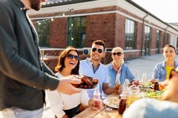leisure and people concept - happy party host offering meat to his friends at barbecue party on rooftop in summer. friends at barbecue party on rooftop in summer