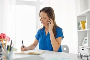 technology, business and people concept - middle-aged woman calling on smartphone and writing to notebook at office or home. woman with notepad calling on smartphone at office
