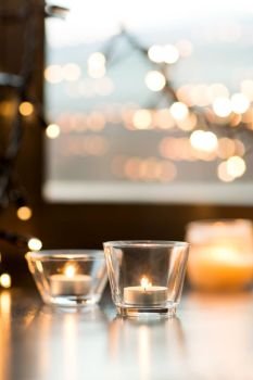 hygge, decoration and christmas concept - candles burning in lanterns and festive garland on window sill at home. candles burning on window sill with garland lights