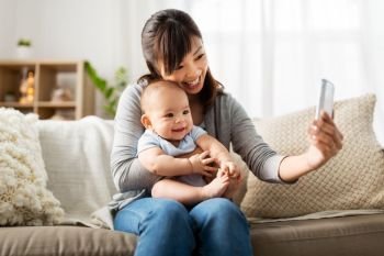 family, technology and motherhood concept - happy young asian mother with little baby son taking selfie by smartphone at home. happy mother with baby son taking selfie at home