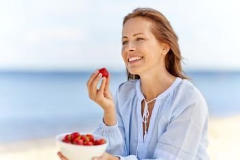 people and leisure concept - happy smiling woman eating strawberries on summer beach. happy woman eating strawberries on summer beach