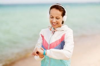 sport, technology and healthy lifestyle concept - smiling woman with fitness tracker and headphones on beach. woman with fitness tracker and headphones on beach