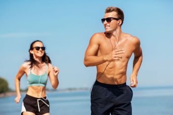 fitness, sport and technology concept - happy couple with earphones running along summer beach. couple with earphones running along on beach
