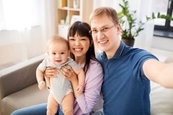 mixed race family, parenthood and people concept - happy mother and father with baby son taking selfie at home. mixed race family with baby taking selfie at home