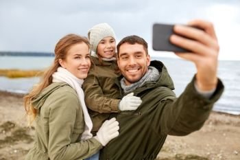 family, leisure and people concept - happy mother, father and little daughter taking selfie by smartphone on autumn beach. family taking selfie by smartphone on autumn beach