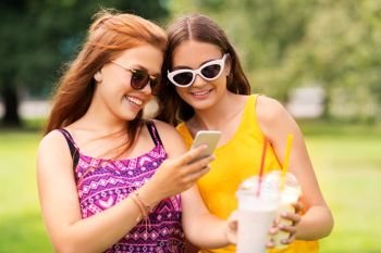 leisure, technology and internet addiction concept - happy smiling teenage girls or friends with smartphone drinking milk shakes or smoothie at summer park. teenage girls with smartphone and shakes in park