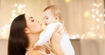 family and motherhood concept - happy smiling young mother kissing little baby over christmas lights background. mother kissing baby over christmas lights