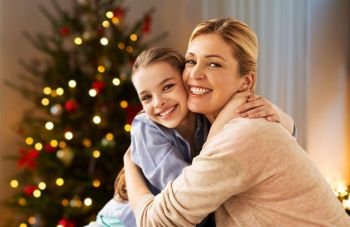people, holidays and family concept - happy smiling girl with mother hugging on sofa at home over christmas tree lights background. happy smiling family hugging at home on christmas