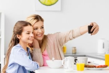 family, technology and people concept - happy mother and daughter with smartphone having breakfast and taking selfie at home kitchen. family taking selfie by smartphone at breakfast