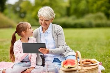 family, leisure and technology concept - happy grandmother and granddaughter with tablet pc computer having picnic at summer park. grandmother and granddaughter with tablet at park