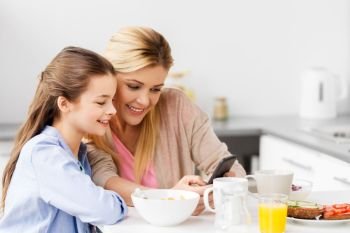 family, technology and people concept - happy mother and daughter with smartphone having breakfast at home kitchen. family with smartphone having breakfast at home