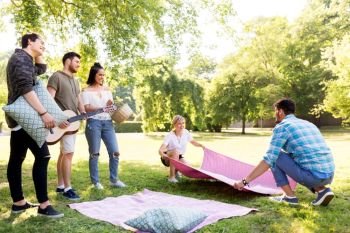 friendship and leisure concept - group of happy friends with guitar, basket and blanket arranging place for picnic at summer park. friends arranging place for picnic at summer park