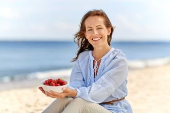 people and leisure concept - happy smiling woman holding bowl with strawberries on summer beach. woman holding bowl with strawberries on beach