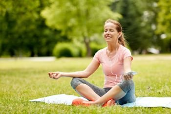 fitness, sport and technology concept - woman with smartphone and earphones meditating on yoga mat at summer park. woman with smartphone and hones meditating at park