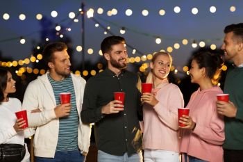 leisure, celebration and people concept - happy friends with drinks at rooftop party at night with blurred bokeh lights. friends with party cups on rooftop at night