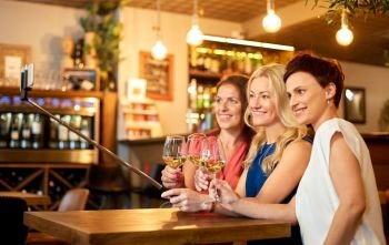 people, technology and lifestyle concept - women drinking wine and taking picture by smartphone on selfie stick at bar or restaurant. women taking picture by selfie stick at wine bar