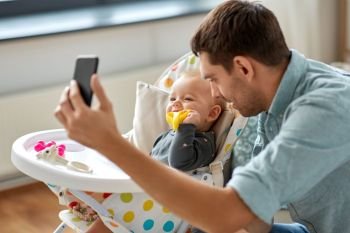 family, fatherhood and people concept - father with little baby daughter in highchair taking selfie by smartphone at home. father with baby daughter taking selfie at home