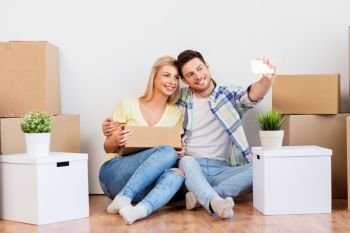 moving, people and real estate concept - smiling couple with big cardboard boxes taking selfie by smartphone at new home. couple taking selfie and moving to new home